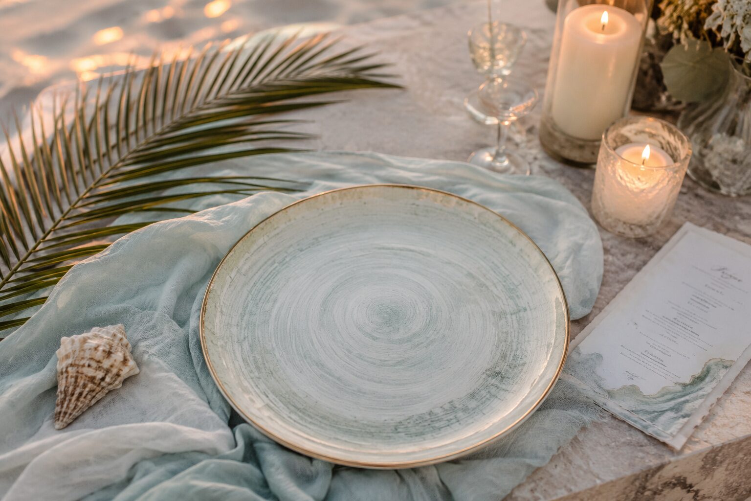 A long table set on the sand at sunset, with tropical flowers, candles and grilled island fare